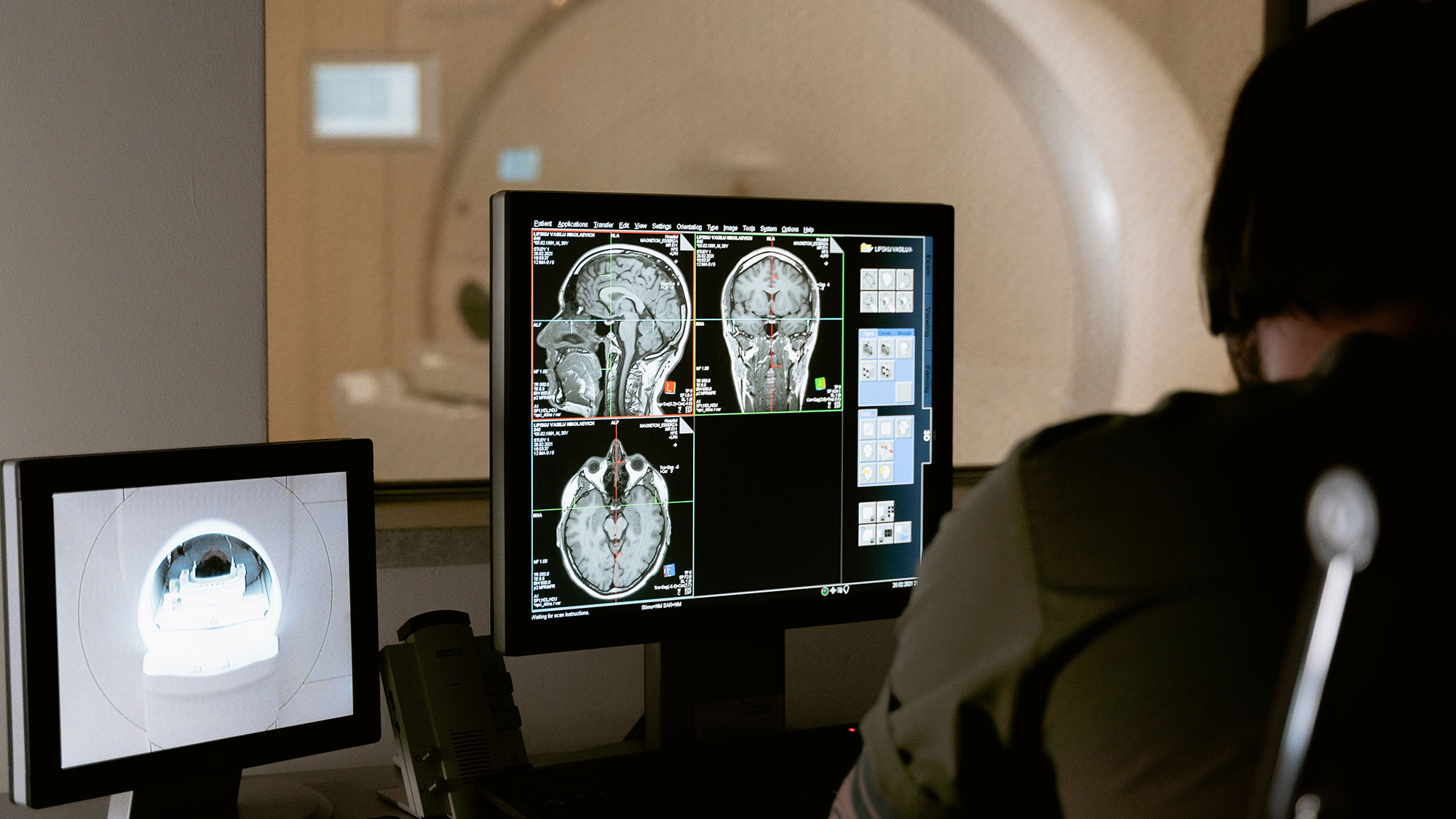 The camera look over the shoulder of a person looking at a computer screen with three brain scans on the monitor that show lit up areas of the brains where activity is occurring.