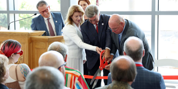 Three people use giant scissors to cut a red ribbon while an audience watches.