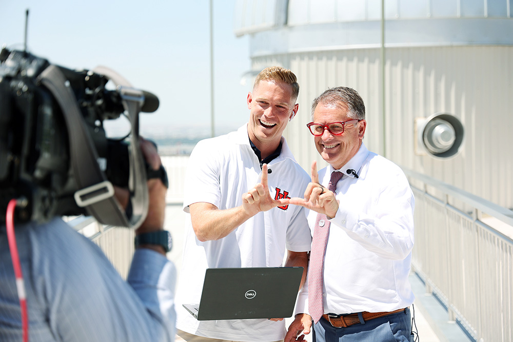 Two men form the letter U with their thumbs and pointer fingers in front of a camera on a rooftop.