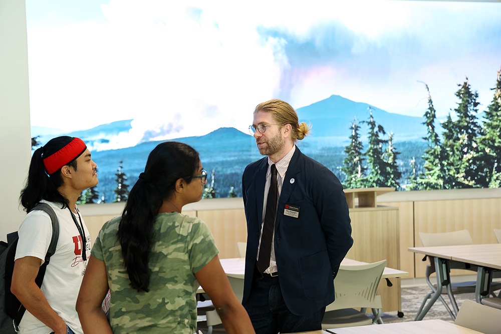 A man stands in front of a giant monitor while talking to two women.