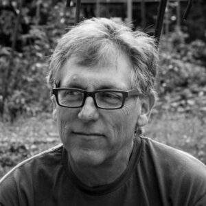 A black and white headshot of a man with black frame glasses taken outside in nature.