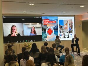 An audience watches a panel of two people on a video screen and two people sitting at the front of the room