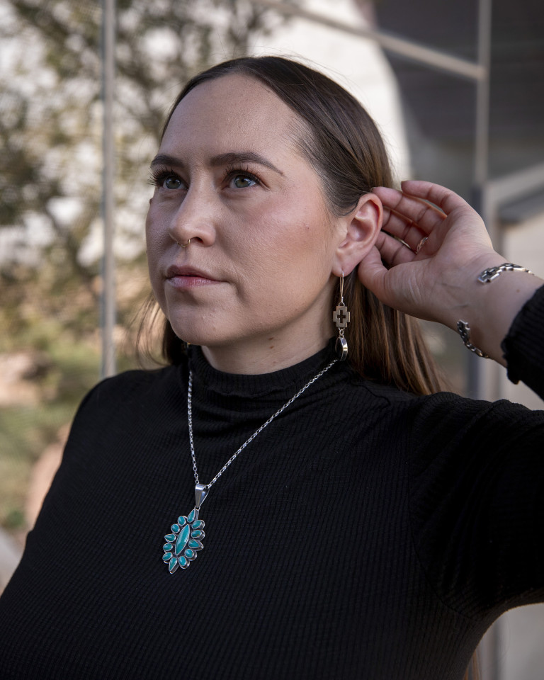A woman in a black mock turtleneck, wearing a silver necklace with turquoise stones and silver earrings with a Navajo cross and a dark oval stone.