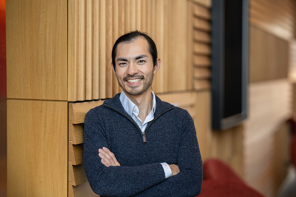 A man poses in a colored sweater, quarter zip, leaning against a textured wall.
