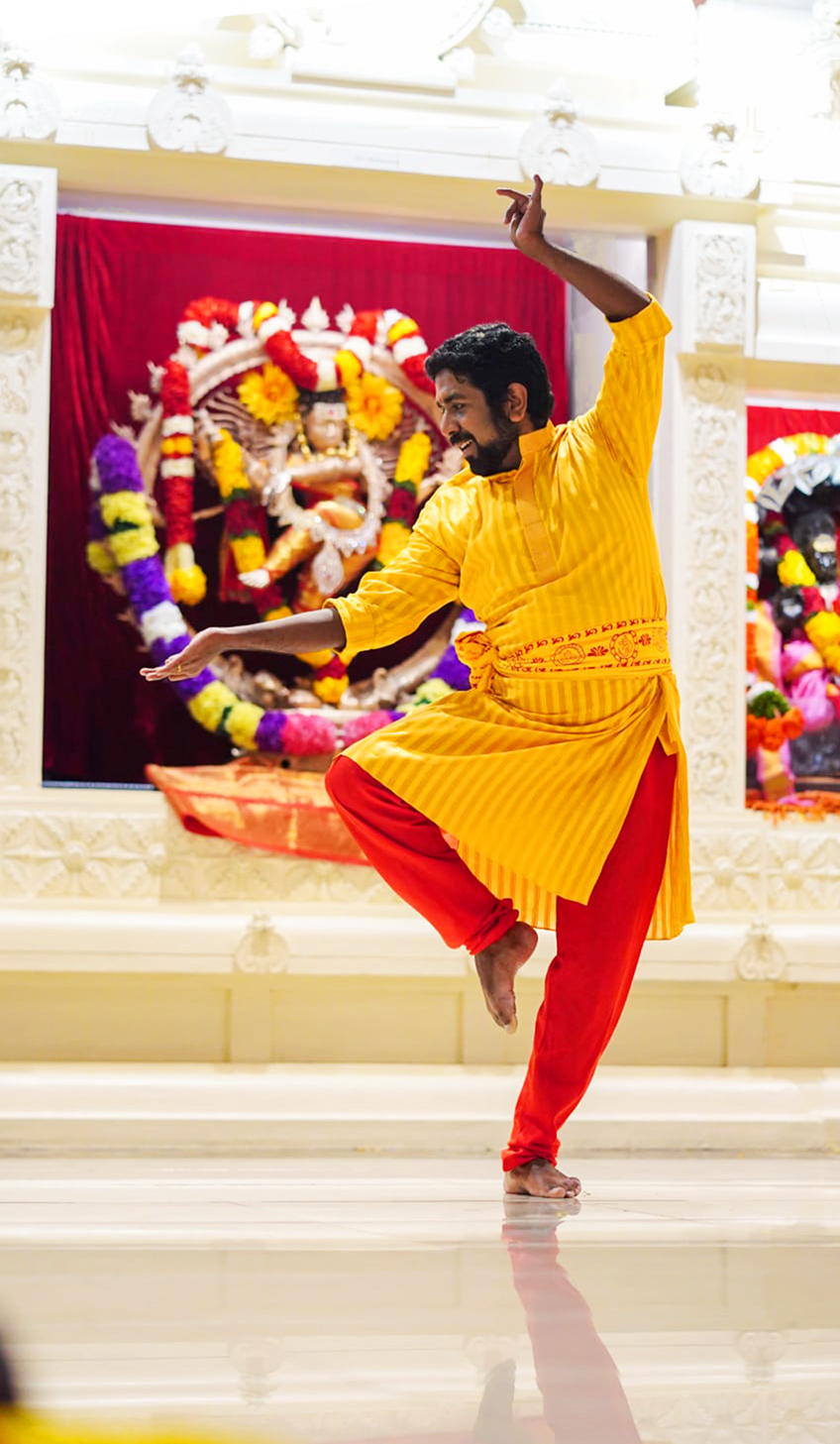 A man is performing a classic Indian dance, wearing a marigold tunic and orange pants. His arms are arranged in a C over his head.