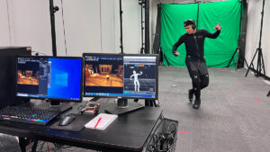 A man dances wearing a spandex motion capture suit, with a person at a computer in the foreground and green screen in the background.