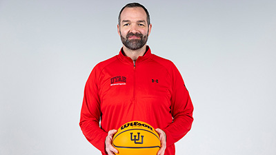 A man in a red U of U jacket holds a basketball and looks at the camera