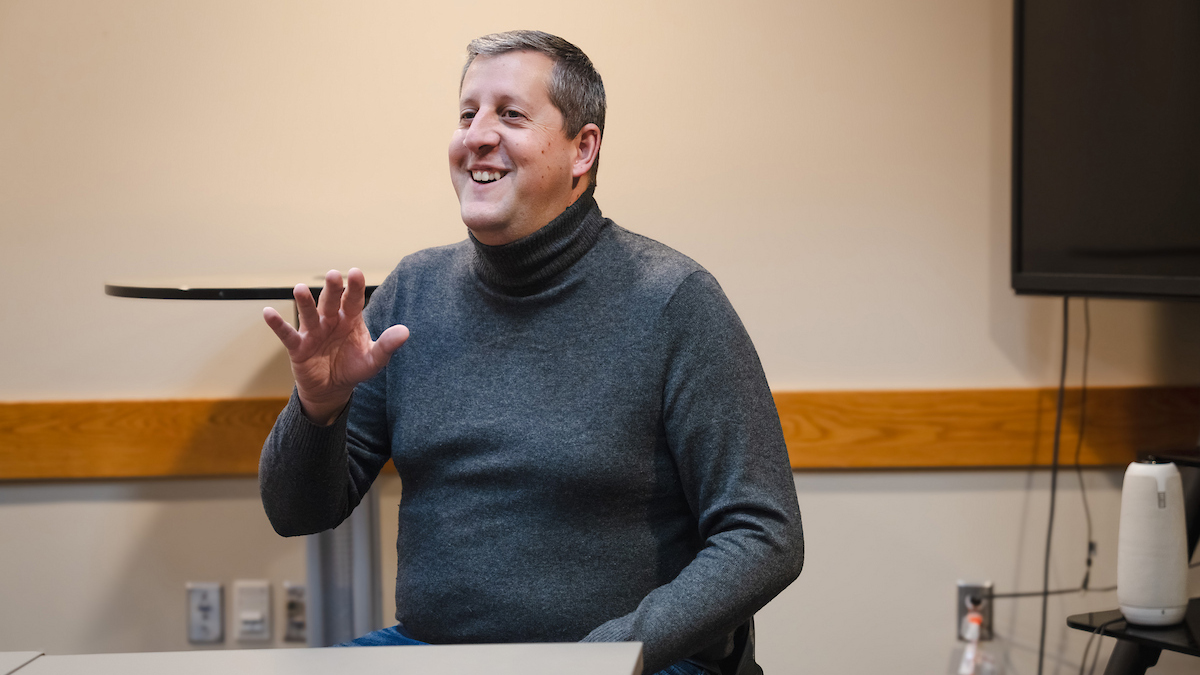 A man smiles while he does American sign language in a gray turtleneck.
