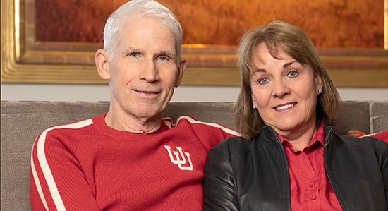 Man with white hair in a red U of U shirt sitting on a couch next to woman with brown hair.