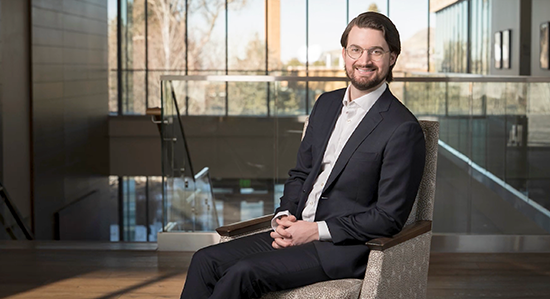Young man with dark hair in a dark suit, leaning back in armchair and smiling.