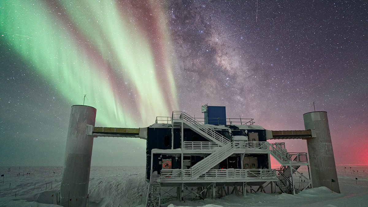 The IceCube Lab is seen under a starry, night sky, with the Milky Way appearing next to striking auroras in the background.