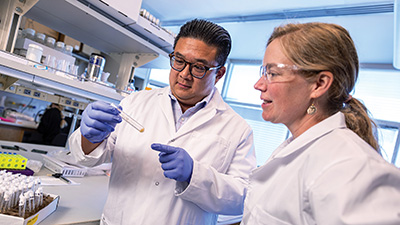 Two University of Utah researchers in a laboratory examining a sample while wearing lab coats and gloves.