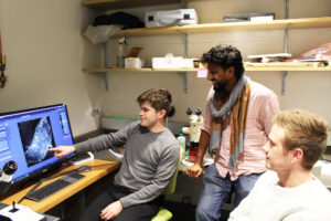 Three men looking at a computer screen-a microscopic image with flurorescent colors. Two are sitting in office chairs and one is leaning against a counter.