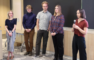 Five people stand in front of a black board, giving a presentation. Left to right is woman man man woman woman, with the left most women speaking into a microphone.