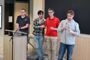 Four men stand to the side of a screen, one man in glasses talking into a microphone.