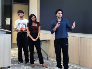 Three people from left to right, man woman man) stand to the side of a projection screen, and one man is talking into a microphone.